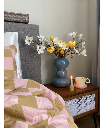 Een nachtkastje met een blauwe vaas met daarin de Kunstbloem Magnolia 50 cm geel/oranje, een roze theekopje op een margrietvormig schoteltje en een brandende kaars staat naast een bed dat is bedekt met een roze en bruine deken met geometrische patronen.