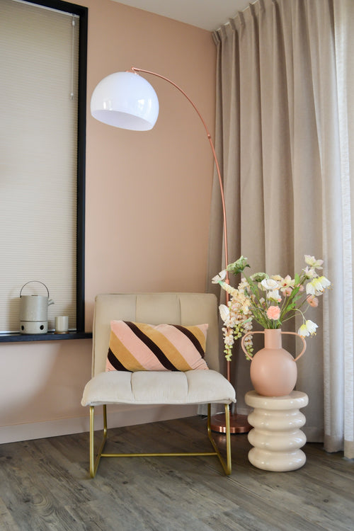 Un angolo moderno e accogliente con una sedia color crema con cuscino a righe, un'alta lampada da terra a forma di arco, tende beige, un pavimento in legno e un vaso rotondo con fiori bianchi artificiali Fritillary 94 cm su un tavolino scolpito.