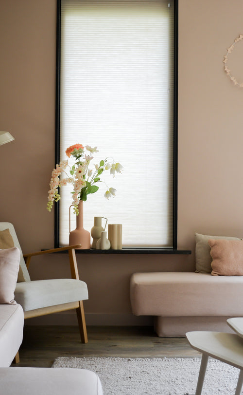 Un accogliente soggiorno con pareti rosa chiaro, una grande finestra con una tenda a rullo bianca, divani color crema e cipria e una sedia in legno con il fiore artificiale Fritillary 94 cm bianco accanto ad oggetti decorativi sul davanzale della finestra.