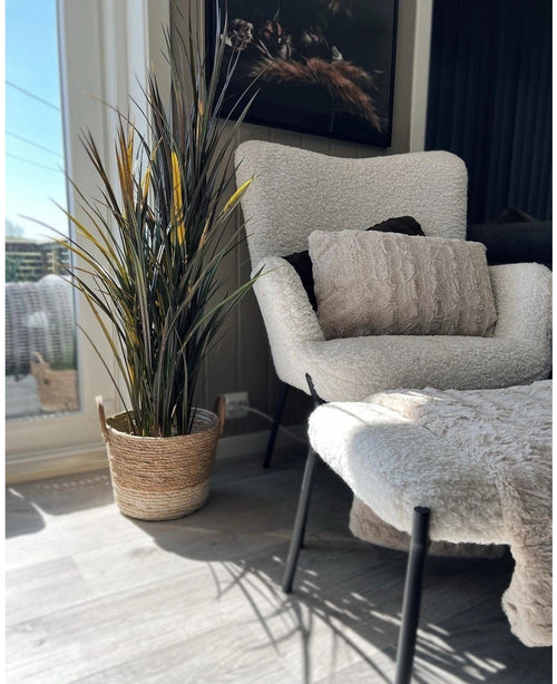 Un angolo accogliente con una soffice poltrona bianca, un pouf coordinato, un cuscino strutturato e l'erba artificiale Bordeaux UV 120 cm in un cestino intrecciato con la luce del sole che splende attraverso una grande finestra.