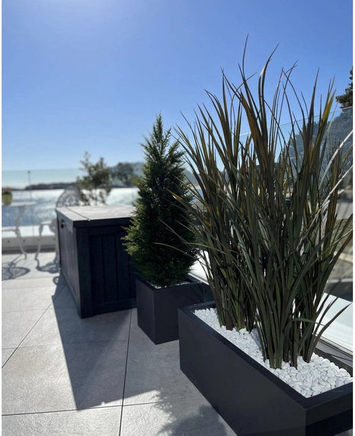 Erba artificiale Bordeaux UV 120 cm in fioriere rettangolari con pietre bianche decora una moderna terrazza all'aperto su uno sfondo di cielo azzurro e alberi in lontananza.