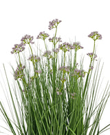 De Kunstgras Prei Bloem Ui Graspot 60cm heeft hoge, dunne groene bladeren en trossen kleine, delicate lichtpaarse bloemen op lange stelen. Het arrangement is brandvertragend en afgebeeld tegen een witte achtergrond.