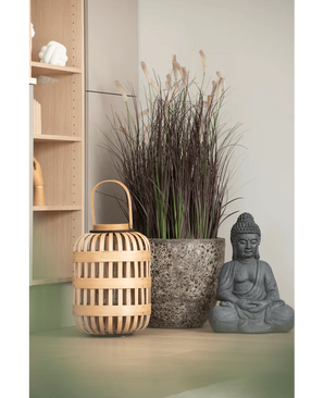 Una lanterna di legno, un alto vaso di pietra con bonsai di erba di cipolla artificiale di 100 cm (ignifugo) e una statua di Buddha grigia sono disposti su una superficie di colore chiaro, vicino a una mensola di legno e a un muro chiaro.