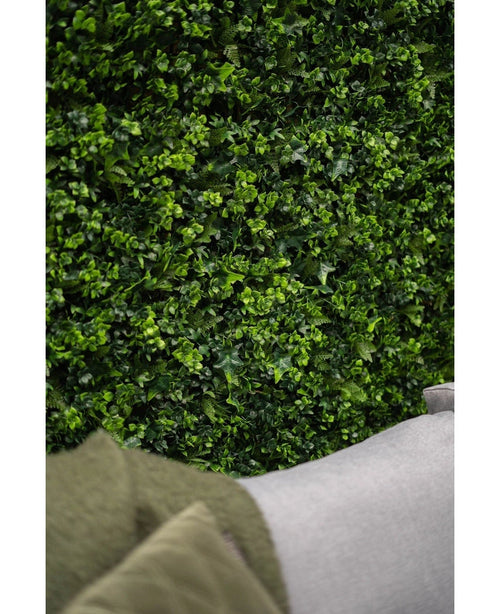 Primo piano di un muro di una siepe artificiale verde lussureggiante mix 50x50 cm con foglie vivaci, con l'angolo di un divano grigio chiaro e parte di un cappotto verde visibile in primo piano in basso.