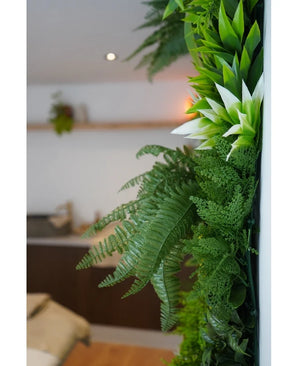 Un primo piano di piante artificiali UV Green Hedge Freshness 100x100 cm verde lussureggiante adorna una parete interna verticale, con uno sfondo sfocato che mostra mobili neutri e una piccola pianta sospesa.