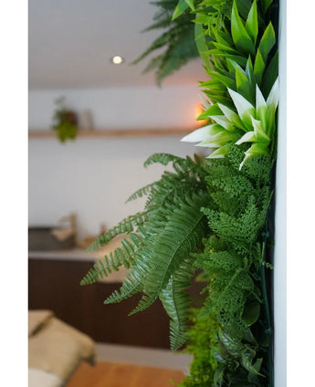 Un primo piano di piante artificiali UV verde lussureggiante di Hedge Freshness 100x100 cm adorna una parete interna verticale, con uno sfondo sfocato che mostra mobili neutri e una piccola pianta sospesa.
