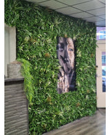 Een Kunsthaag Savanne brandvertragend paneel van 100x100 cm met weelderig groen omringt een groot zwart-wit gezichtsportret in een binnenruimte met betegelde plafondpanelen en een kleine toonbank aan de linkerkant.