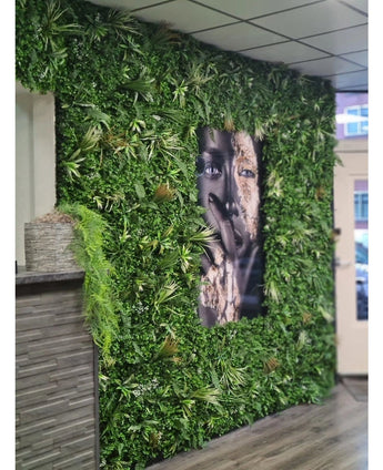 Een Kunsthaag Savanne brandvertragend paneel van 100x100 cm met weelderig groen omringt een groot zwart-wit gezichtsportret in een binnenruimte met betegelde plafondpanelen en een kleine toonbank aan de linkerkant.