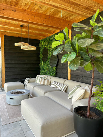 Een gezellige zithoek buiten met een beige sofa, gestreepte kussens, een ronde vuurplaats, een grote plant in potten en een houten pergola. De muur is voorzien van groen met de Kunsthaag Volta 100x100 cm UV voor extra stijl.