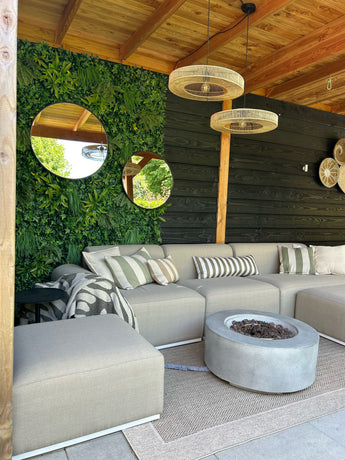 Een gezellig terras met een beige sofa, gestreepte kussens, vuurplaats en geweven vloerkleden. Aan de muur hangt UV-groen van Kunsthaag Volta 100x100 cm, ronde spiegels en rieten lampen onder een houten pergola.