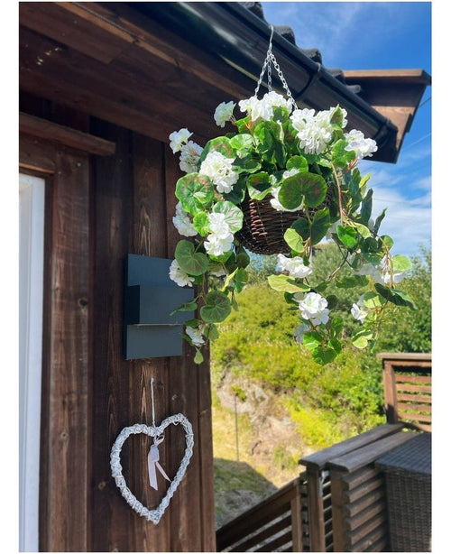 La Begonia artificiale bianca in cesto UV 70 cm è appesa a una parete di legno, con un cuore bianco decorativo sotto. Il verde, una terrazza in legno e un cielo azzurro parzialmente nuvoloso completano lo sfondo.