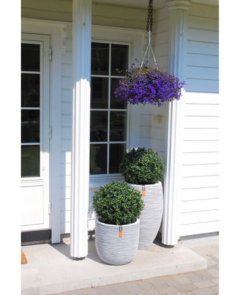 La veranda bianca di una casa ha due fioriere di bosso artificiale su stelo in stile romano UV da 65 cm con arbusti verdi, un cesto sospeso con fiori viola sopra, colonne bianche e una porta con pannelli di vetro sulla sinistra.