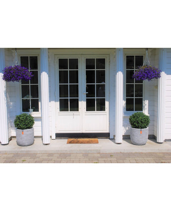Le doppie porte bianche con vetri sono fiancheggiate da due arbusti artificiali UV da 65 cm in bosso artificiale su stelo in stile romano in grandi vasi. Due cesti appesi con fiori viola e uno zerbino completano l'ingresso.