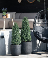 Twee Kunstplant Buxus Pyramide UV 120 cm staan op een houten dek voor een buitenzitje met een lichte bank, ronde tafel met lavendel en zwarte hangstoel.