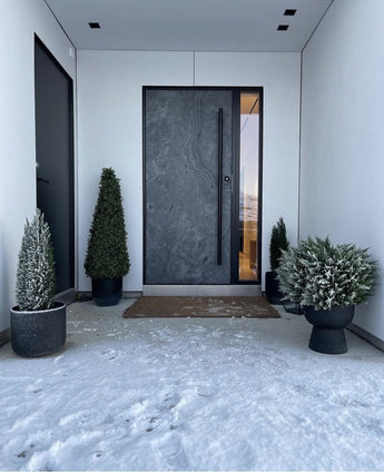 Een moderne donkere voordeur wordt omlijst door witte muren en geflankeerd door Kunstplant Buxus Pyramide UV 90 cm, die UV-bescherming biedt. Lichte sneeuw bedekt de grond, terwijl deze stijlvolle tuinplanten een opgeruimde, minimalistische ingang creëren.