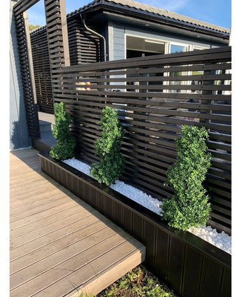 Drie Kunst Buxus Spiraal Romeinse stijl UV 90cm vormsnoeistruiken staan in een smal bed van witte kiezels, tegen een modern zwart privacyhek met latten naast een houten terras en een eigentijds huis.