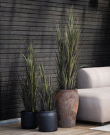 Drie Kunstgras Bordeaux UV 150 cm kunstplanten van verschillende hoogtes staan naast een lichtgekleurde bank op een houten vloer, met op de achtergrond een wand van donker hout.