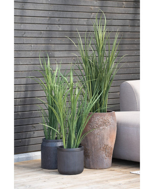 Una pianta artificiale Artificial Grass Green UV da 90 cm in un vaso nero si trova su un pavimento di legno accanto a un divano da esterno grigio, con una parete a doghe di legno scuro sullo sfondo.