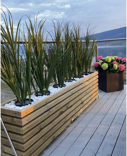 Una fioriera in legno con erba artificiale verde UV 90 cm e pietre bianche si trova su un ponte di legno accanto a una fioriera nera con ortensie colorate, con balaustre in vetro e vista sulle montagne sullo sfondo.