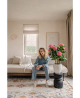 Een persoon met krullend haar zit op een lichtgekleurde bank in een stijlvolle woonkamer met Kunstplant Hortensia UV 85 cm roze, een vloerkleed met patroon, lichte muren en natuurlijk licht dat door het raam naar binnen valt.