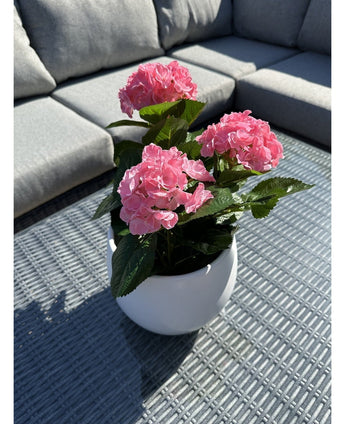 De Kunstplant Hortensia Roze UV 35 cm, een witte pot met levendige roze hortensiabloemen, staat op een geweven grijze buitentafel met een grijze sofa met kussens op de achtergrond, verlicht door zonlicht en zachte schaduwen.
