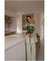 Een vrouw met krullend blond haar in een lichtgroen pak glimlacht terwijl ze de Kunstbloem Kalanchoë plant UV 31 cm wit vasthoudt in een moderne, neutraal getinte keuken met open planken.
