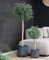 Een kunst olijfboom van 85 cm hoogte (UV-bestendig) staat op een houten patio naast een beige buitenbank voor een donkerhouten wand.