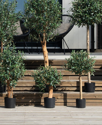Verschillende Kunst olijfbomen 85 cm UV staan op houten treden bij de patio, met zwarte stoelen en een glazen deur erachter. Deze UV-bestendige kunstplanten voegen groen toe aan de ruimte en vragen minimale verzorging.