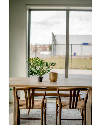 Een houten tafel met twee stoelen, een Kunstpalm Parlour UV 60 cm in een pot en een gouden vaas staan bij een groot raam met uitzicht op een industriegebied met gebouwen en een hek.