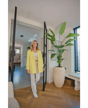 Een glimlachende vrouw in een gele blazer staat in een lichte, moderne kamer met houten vloeren, naast een Kunstplant Strelitzia 210 cm real touch bij de glazen deur. Zonlicht filtert door de jaloezieën aan de rechterkant.