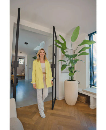 Een glimlachende vrouw in een gele blazer staat in een lichte, moderne kamer met houten vloeren, naast een Kunstplant Strelitzia 210 cm real touch bij de glazen deur. Zonlicht filtert door de jaloezieën aan de rechterkant.