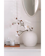 Een Kunstmagnoliatak Real Touch Wit 98cm staat in een ronde witte vaas op een wit oppervlak naast een brandende glazen kaars en een klein flesje op een houten dienblad, in een lichte, minimale kamer met een ronde spiegel erboven.