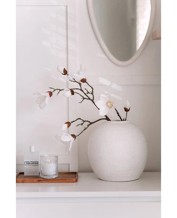 Een Kunstmagnoliatak Real Touch Wit 98cm staat in een ronde witte vaas op een wit oppervlak naast een brandende glazen kaars en een klein flesje op een houten dienblad, in een lichte, minimale kamer met een ronde spiegel erboven.