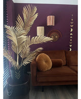 Een moderne woonkamer met een bruin fluwelen bank, rond plooikussen en de Kunstpalm Areca goud 120 cm in een zwarte vaas als gouddecor. Geometrische wandkunst hangt op een donkerpaarse muur, met houten vloeren en verticale lamellen in het zicht.