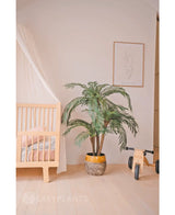 Een gezellige kinderkamer met een licht houten ledikant, de Kunstpalm Cycas Classic 135 cm in een gele plantenbak, een houten speelgoedfiets met het label MIA, minimalistische lijnkunst aan de muur, lichthouten vloeren en zachtroze beddengoed voor extra warmte.