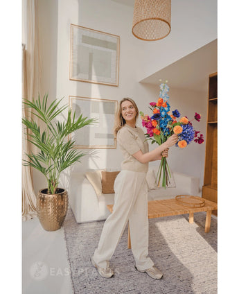 Een glimlachende vrouw in lichtbeige kleding en sneakers staat in een lichte, moderne woonkamer met houten meubels en ingelijste kunst, en houdt een kleurrijk boeket vast. De Kunstpalm Kentia 135 cm voegt een tropisch tintje toe aan de ruimte.