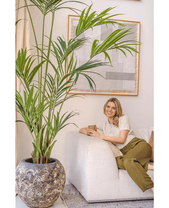 Een vrouw met blond haar, in een crèmekleurige top en olijfgroene broek, zit glimlachend op een witte bank en houdt een mok vast. De Kunstpalm Kentia 220 cm versterkt de tropische sfeer van de lichte, gezellige kamer met abstracte wandkunst.