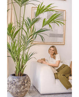 Een vrouw met blond haar, in een crèmekleurige top en olijfgroene broek, zit glimlachend op een witte bank en houdt een mok vast. De Kunstpalm Kentia 220 cm versterkt de tropische sfeer van de lichte, gezellige kamer met abstracte wandkunst.