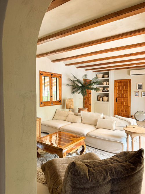 Un accogliente soggiorno ha un grande divano color crema, tavolino da caffè in legno, palma artificiale Phoenix De Luxe da 210 cm in vaso, soffitto con travi in ​​legno, scaffali incorporati con ceramiche, luce naturale dalla finestra e un arredamento caldo e rustico.
