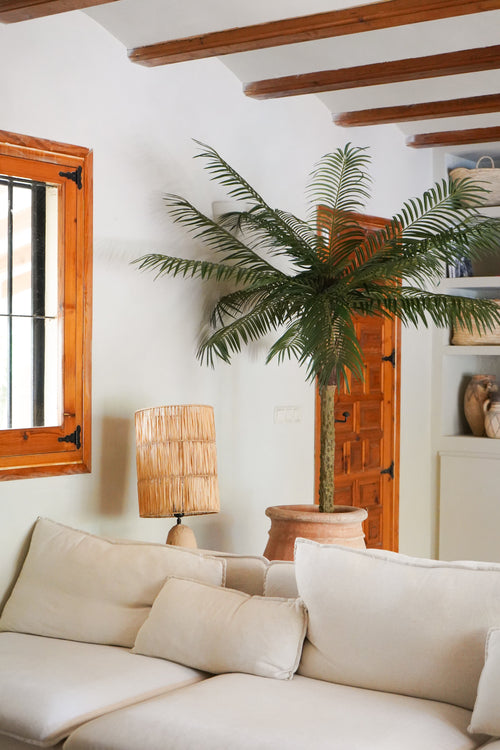 Un accogliente soggiorno con un divano bianco, una lampada da tavolo in rattan, la palma artificiale Phoenix De Luxe da 210 cm in un vaso, travi in ​​legno sul soffitto, una finestra con cornici in legno e una porta in legno sullo sfondo.