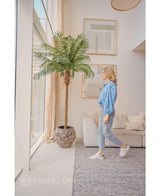 Een vrouw in een blauwe blouse en spijkerbroek loopt langs de Kunstpalm Phoenix De Luxe 250 cm in een lichte, moderne woonkamer met hoge ramen, neutrale inrichting en ingelijste kunst aan de muur.