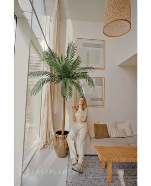 Una donna vestita di colori chiari sta accanto alla Palma artificiale Phoenix De Luxe 250 cm in un soggiorno moderno e luminoso con grandi finestre, un divano beige, una lampada in vimini, un tavolino in legno e quadri murali incorniciati.