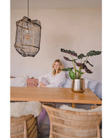 Een vrouw in een witte blouse zit aan een houten eettafel met rieten stoelen. De gezellige, zacht verlichte kamer heeft de Kunstplant Alocasia UV 67 cm en een hanglamp met textuur als decor.