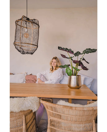 Een vrouw in een witte blouse zit aan een houten eettafel met rieten stoelen. De gezellige, zacht verlichte kamer heeft de Kunstplant Alocasia UV 67 cm en een hanglamp met textuur als decor.