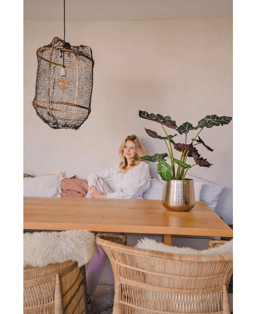 Una donna con una camicetta bianca siede a un tavolo da pranzo in legno con sedie di vimini. La stanza accogliente e delicatamente illuminata ha come decorazione la pianta artificiale Alocasia UV 67 cm e una lampada a sospensione strutturata.