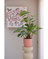 De Kunstplant Appelblad 90 cm, een groenbladige potplant in een roze geribbelde pot, staat op een beige sokkel voor een muur met abstracte roze bloemsierkunst en een doorschijnend wit gordijn aan de rechterkant.