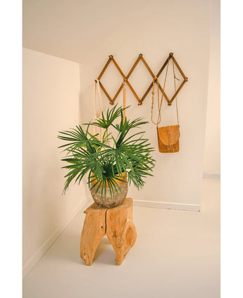 De Kunstplant Baby Palm Fan in pot UV 50 cm zit op een houten kruk in een minimalistische kamer. Daarachter toont een bruin wandrek in accordeonstijl bruine en bruine leren tassen tegen een kraakheldere witte muur.