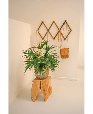 De Kunstplant Baby Palm Fan in pot UV 50 cm zit op een houten kruk in een minimalistische kamer. Daarachter toont een bruin wandrek in accordeonstijl bruine en bruine leren tassen tegen een kraakheldere witte muur.