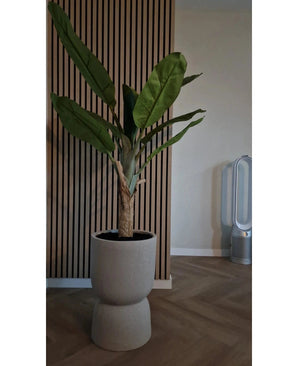 De Kunstplant Bananenboom 150 cm met grote groene bladeren staat op een houten vloer voor een houten wand met latten. Rechts staat een moderne bladloze ventilator in de kamer met neutrale muren en vloer.