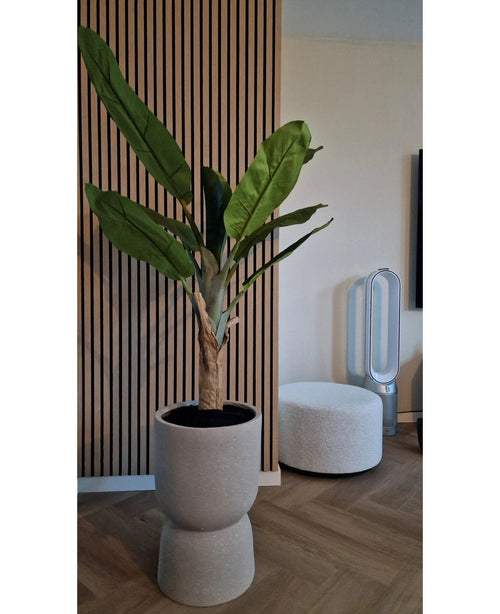 Il Banano Artificiale 150 cm, con le sue foglie verdi lussureggianti, si erge su un pavimento di legno vicino ad una parete nervata con pannelli di legno. Nelle vicinanze c'è un pouf rotondo bianco e un ventilatore senza pale sullo sfondo.